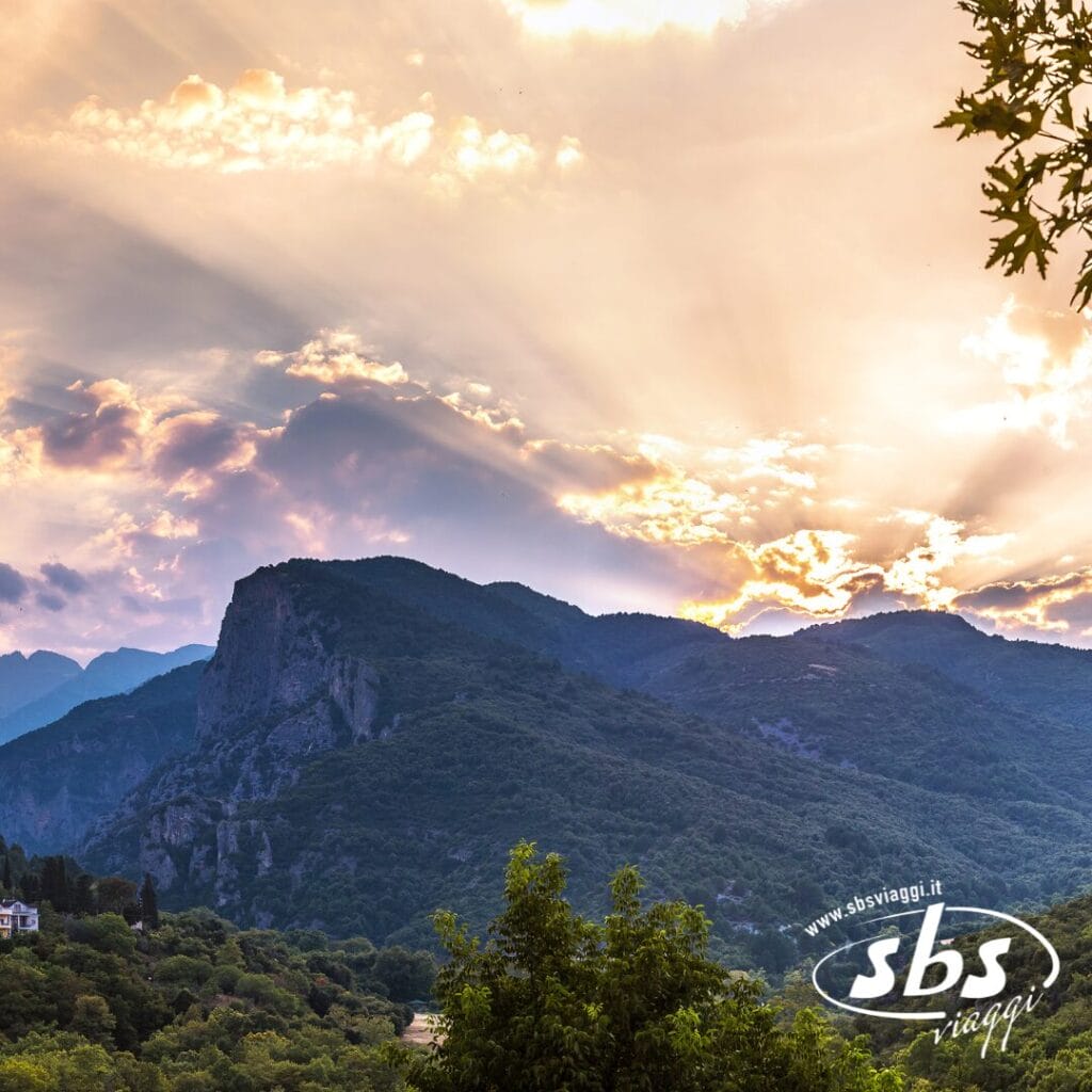La luce del sole filtra tra le nuvole su un paesaggio montuoso dalla vegetazione lussureggiante, evocando la bellezza delle Terre di Alessandro Magno. Il logo