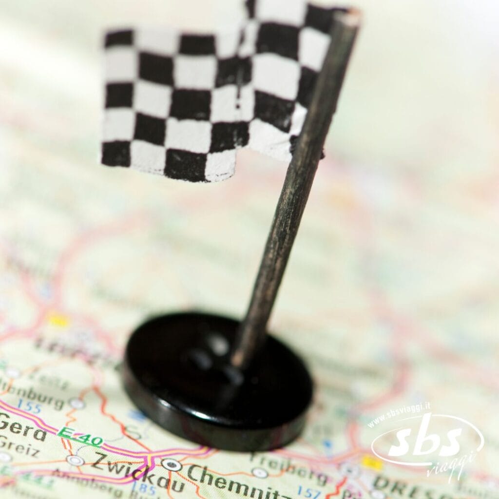 Una piccola spilla a forma di bandiera a scacchi indica un punto sulla mappa, con città e strade visibili sullo sfondo: perfetta per seguire il Grand Tour del 2025 o pianificare le tue avventure in MotoGP.