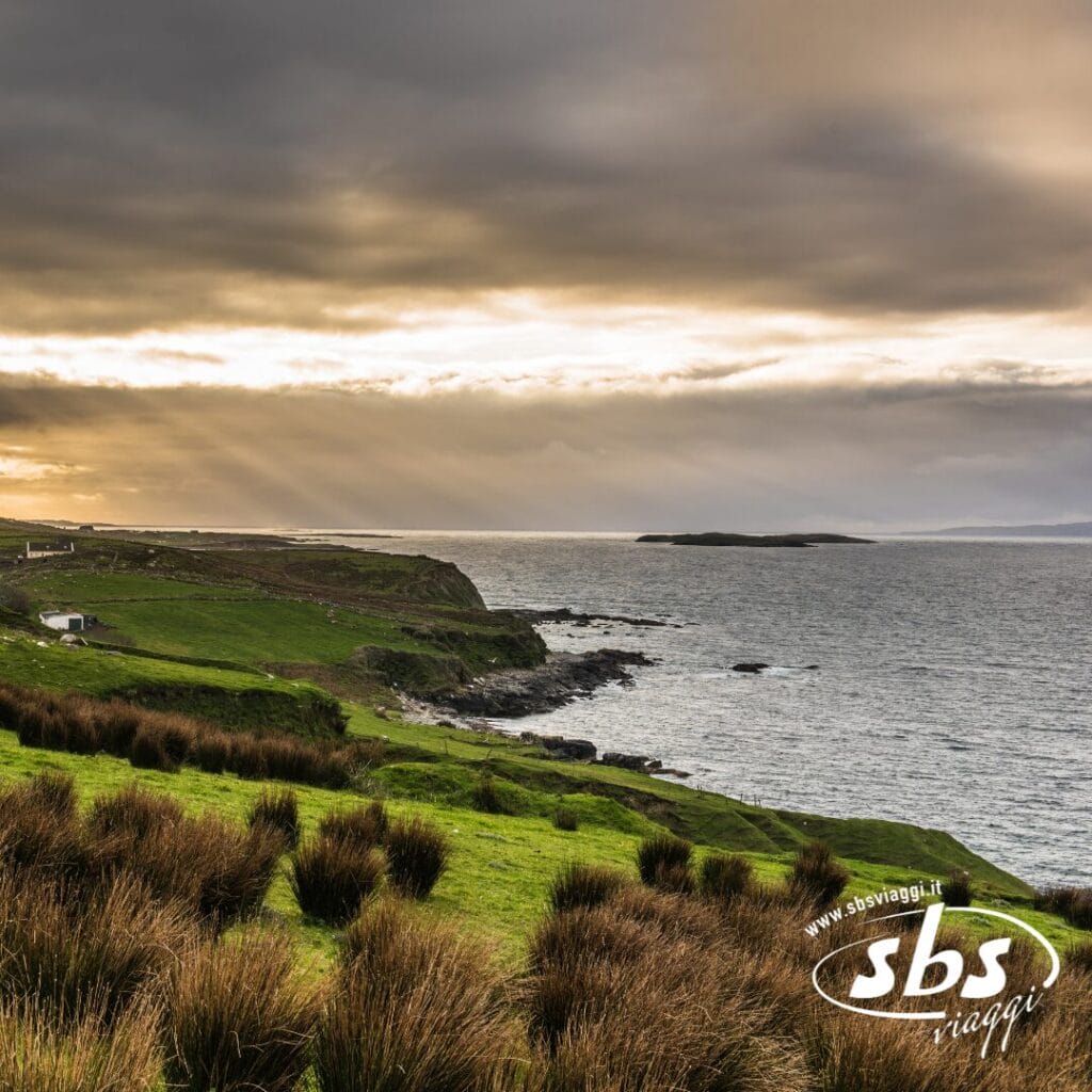 Paesaggio costiero con scogliere erbose verdi, costa rocciosa e cielo nuvoloso con raggi di sole che filtrano. Godetevi il panorama con un cocktail irlandese in mano. Logo "sbs viaggi" in basso a destra.