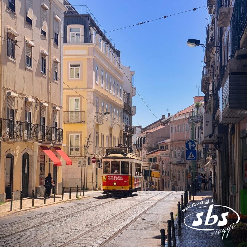 Un tram giallo percorre una strada acciottolata fiancheggiata da edifici color pastello sotto un cielo azzurro e terso in una città europea, catturando l'essenza del Portogallo Autentico. Il logo