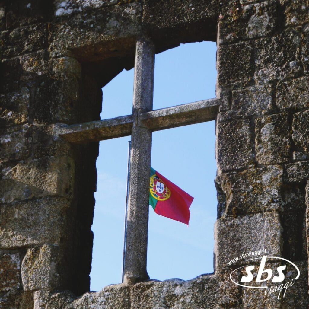 Una bandiera portoghese è visibile attraverso la cornice in pietra di una finestra di un vecchio edificio, evocando lo spirito vibrante di un Cocktail Portoghese. Il cielo è limpido e azzurro sullo sfondo.