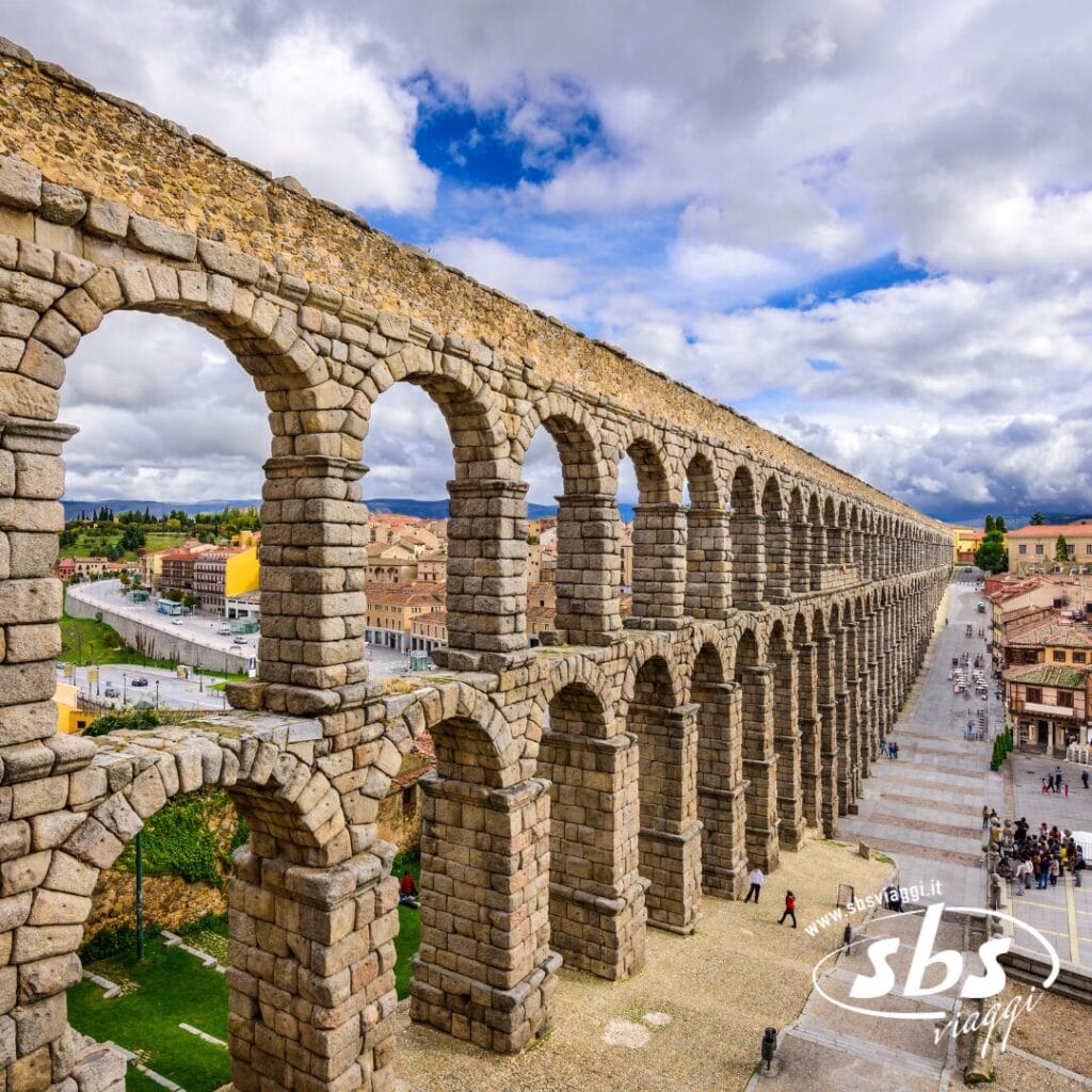 L'antico acquedotto in pietra, una vera meraviglia di architettura, si estende con i suoi molteplici archi attraverso il paesaggio urbano sotto un cielo parzialmente nuvoloso. Come parte del tour Spagna Medievale, i visitatori possono passeggiare sotto questa magnifica struttura, ammirandone la storica grandiosità.