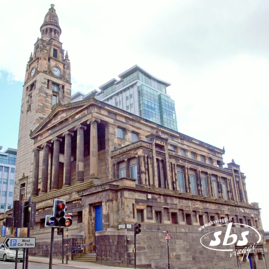 Uno storico edificio in pietra con colonne e una torre dell'orologio si erge accanto a moderni edifici in vetro presso un incrocio cittadino con semafori, offrendo uno spettacolo indimenticabile a chiunque faccia un tour delle Lofoten.