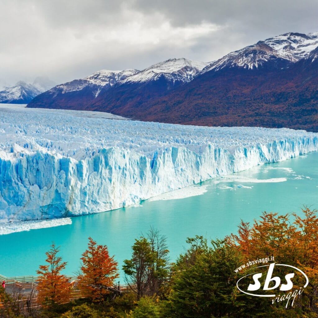 Un grande ghiacciaio incontra l'acqua turchese nel Sudamerica, circondato da alberi dai colori autunnali e montagne innevate sotto un cielo nuvoloso; logo