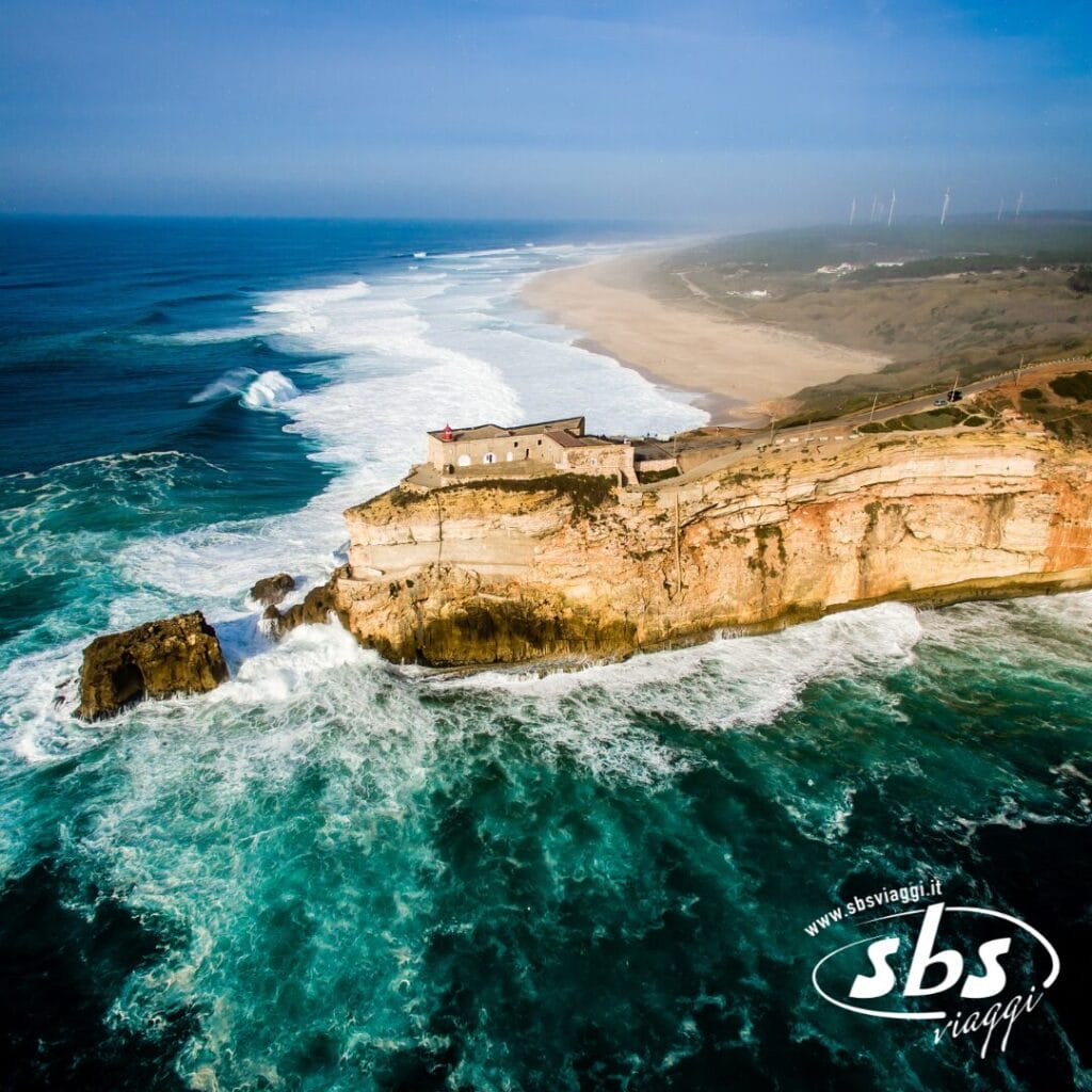 Vista aerea di una scogliera costiera in Portogallo, con un edificio circondato dalle onde che si infrangono e dall'immensità dell'oceano, sotto un cielo azzurro e limpido: una vera meraviglia da esplorare durante il tuo prossimo tour.