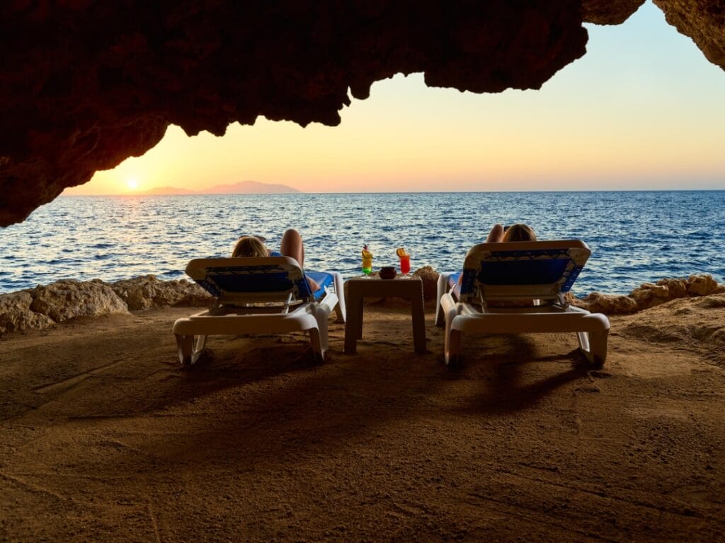 Due persone si rilassano sui lettini prendisole in una grotta serena, di fronte all'oceano al tramonto. I drink sono appoggiati su un tavolino tra loro, creando una scena rinfrescante come una Bozza automatica.