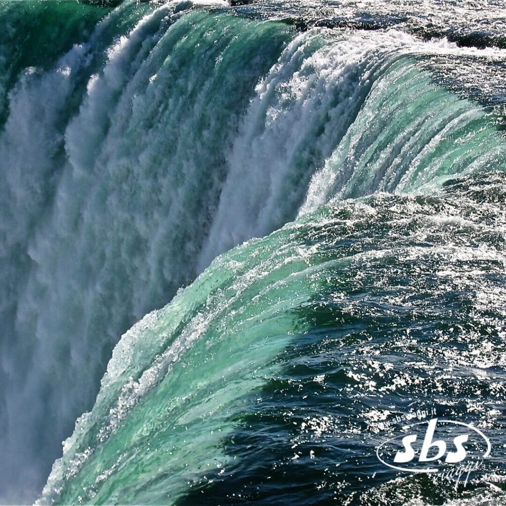 Primo piano dell'acqua che scorre oltre il bordo delle Cascate del Niagara, con la luce del sole che si riflette sulla superficie; nell'angolo in basso a destra è visibile il logo