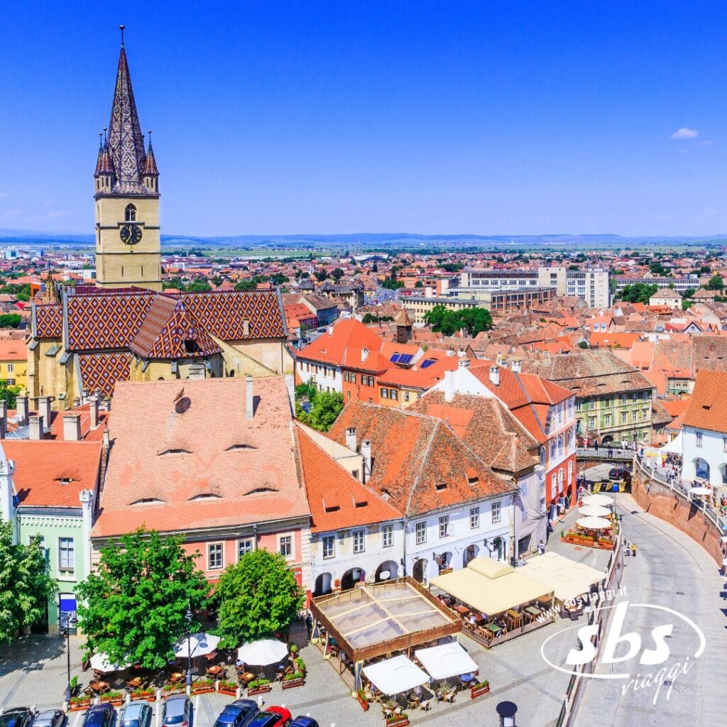 Vista aerea di una piazza cittadina europea in Romania, con caffè all'aperto, edifici dai tetti rossi, una chiesa con tetto decorato e una torre dell'orologio sotto un cielo limpido: perfetta per chi desidera visitare la Romania.