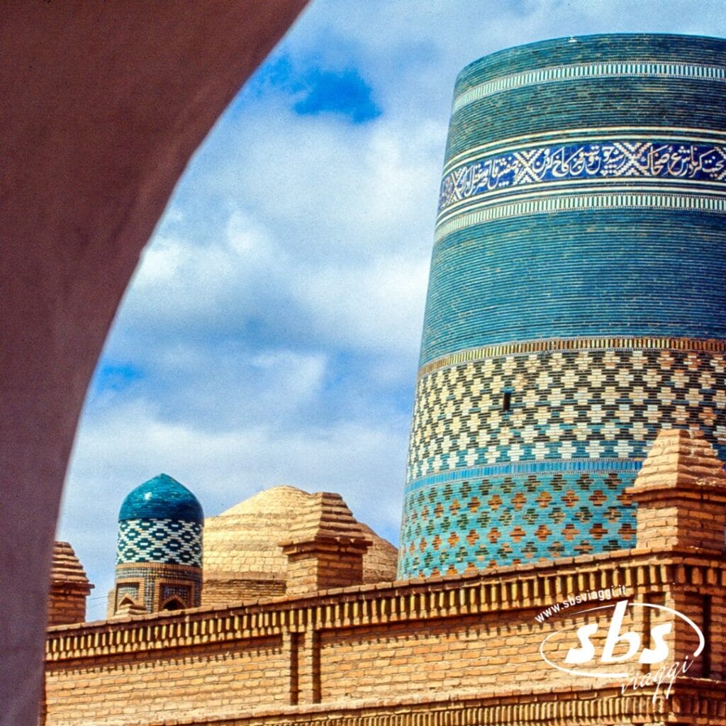 Un minareto e delle cupole ricoperti di piastrelle blu si ergono sopra un muro di mattoni a Khiva, in Uzbekistan, lungo la storica Via Della Seta, con parte di un arco in primo piano e un cielo azzurro sullo sfondo.