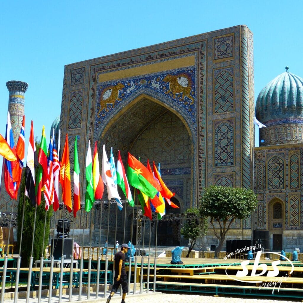 Uno storico edificio architettonico islamico con intricate decorazioni in piastrelle, un grande ingresso ad arco e numerose bandiere nazionali esposte sulla facciata, che ricordano la magnificenza della Terra di Tamerlano.