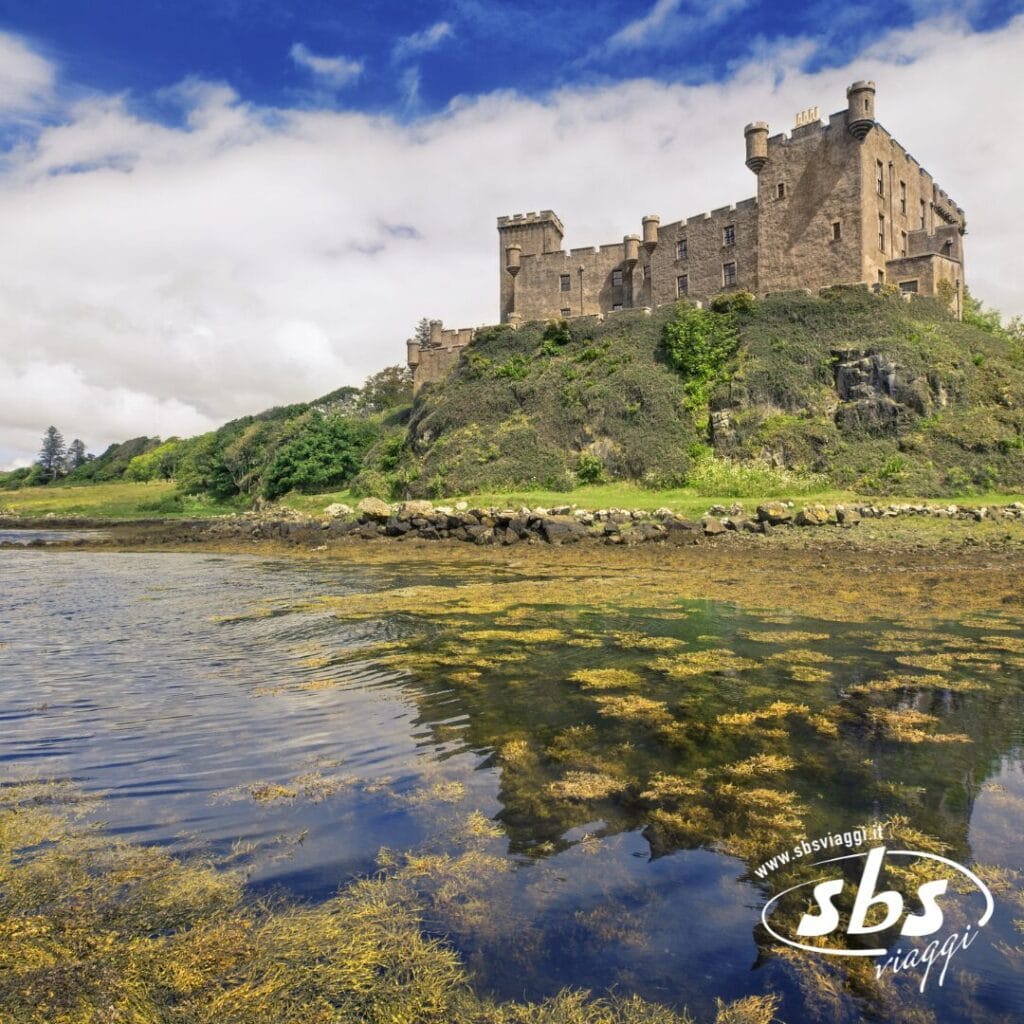 Un classico castello in pietra si erge sulla cima di una collina erbosa che domina un corso d'acqua in Scozia, con cielo azzurro e nuvole sopra; vegetazione e rocce punteggiano la pittoresca costa.