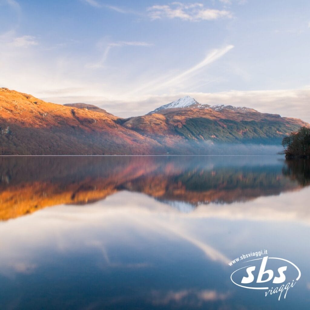 Un lago calmo riflette le colline dai colori autunnali e una montagna innevata sotto un cielo azzurro, catturando la serena bellezza di un viaggio in Scozia; il logo