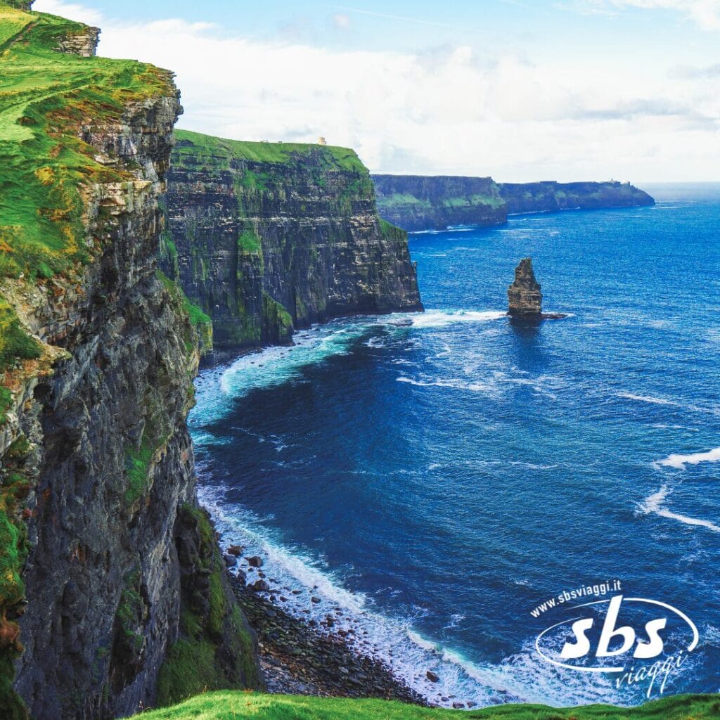 Ripide scogliere verdi si ergono sull'azzurro Oceano Atlantico presso le Scogliere di Moher in Irlanda, con le onde che si infrangono sotto e un faraglione di rocce visibile: una delle vere bellezze d'Irlanda per viaggi indimenticabili.