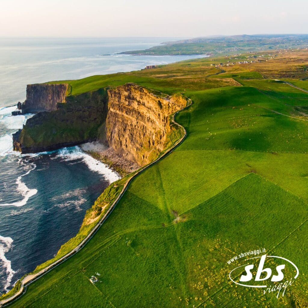 Vista aerea delle ripide scogliere costiere e degli ampi campi verdi che incontrano l'oceano in Irlanda, con una strada tortuosa visibile; il logo