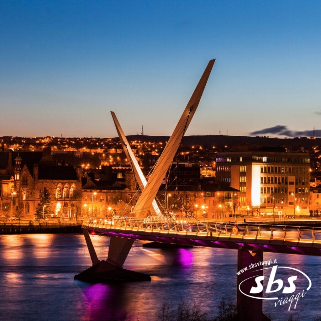 Un moderno ponte pedonale illuminato, con due sostegni angolari, attraversa un fiume al tramonto a Belfast, in Irlanda, con edifici e luci della città che brillano sullo sfondo.