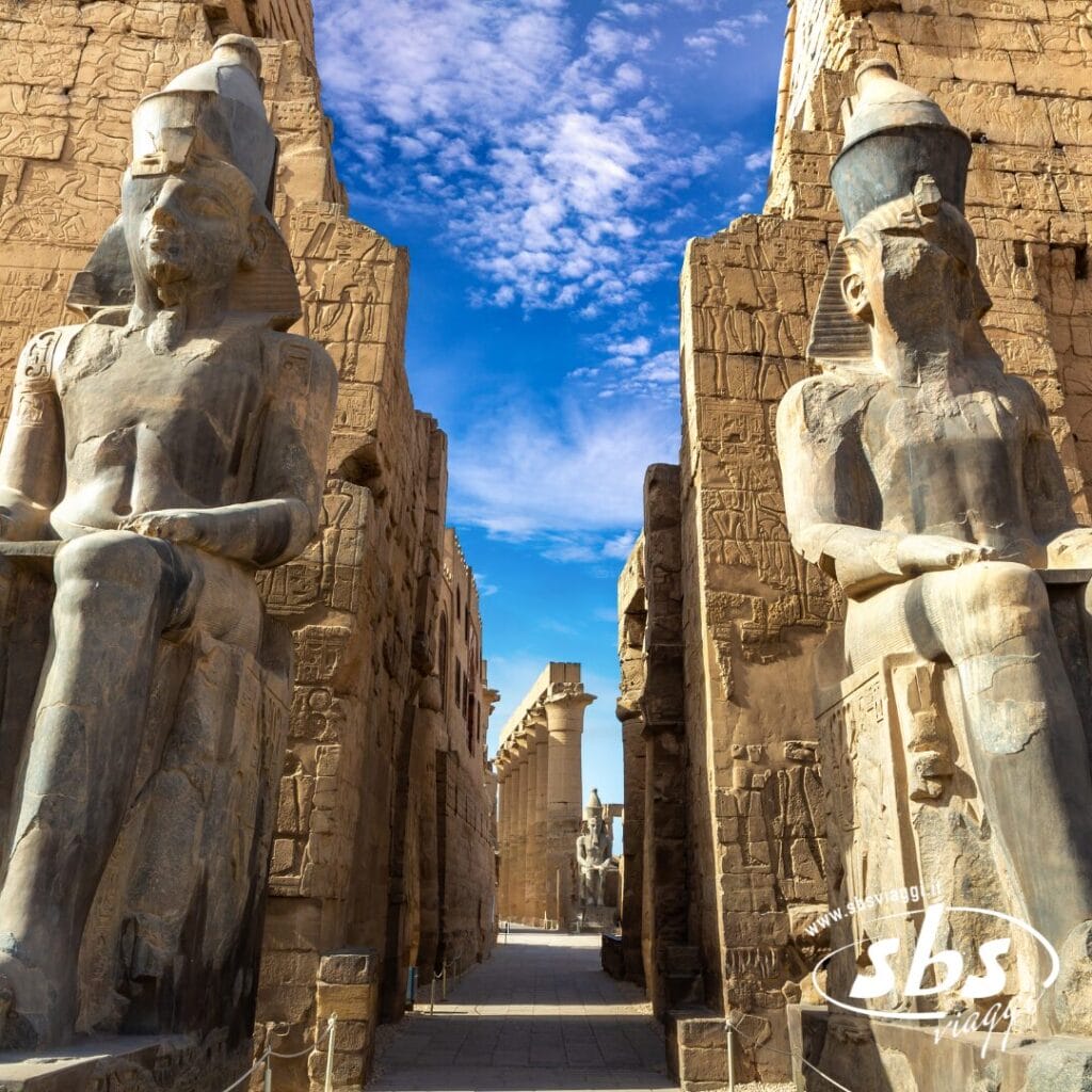 Due grandi statue sedute di antichi faraoni egizi fiancheggiano l'ingresso del tempio, con colonne e pareti in pietra scolpita. Questo sito maestoso ricorda le antiche meraviglie lungo il Nilo, sotto un cielo azzurro con nuvole sparse.