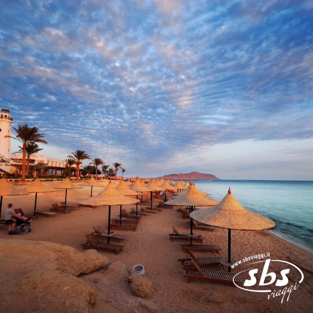 Spiaggia con file di sedie a sdraio vuote e ombrelloni di paglia al tramonto, incorniciata da palme, un faro e montagne in lontananza, che evocano la tranquilla atmosfera di Sharm sul Nilo.