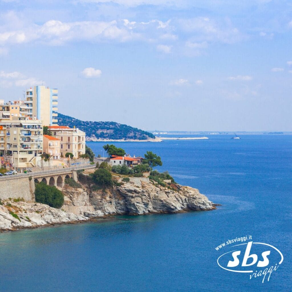 Paesaggio urbano costiero con edifici a picco sul mare azzurro sotto un cielo parzialmente nuvoloso; scopri Salonicco, il gioiello delle Terre di Alessandro Magno, nel tuo prossimo tour. Logo SBS Viaggi nell'angolo.