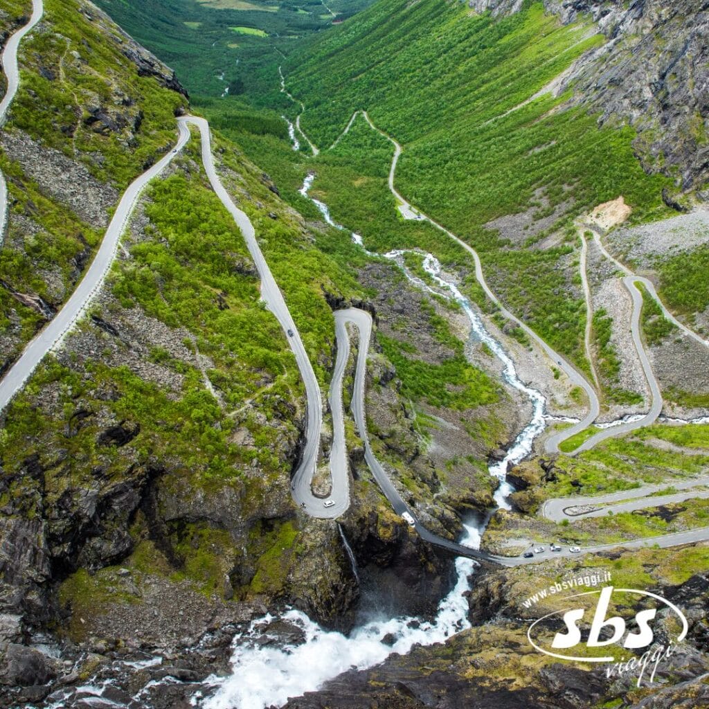 Una ripida e tortuosa strada di montagna con numerosi tornanti stretti attraversa terreni rocciosi e verdi colline, offrendo uno spettacolo naturale con un fiume e una cascata sottostanti, che ricordano il suggestivo Fiordi Norvegesi.