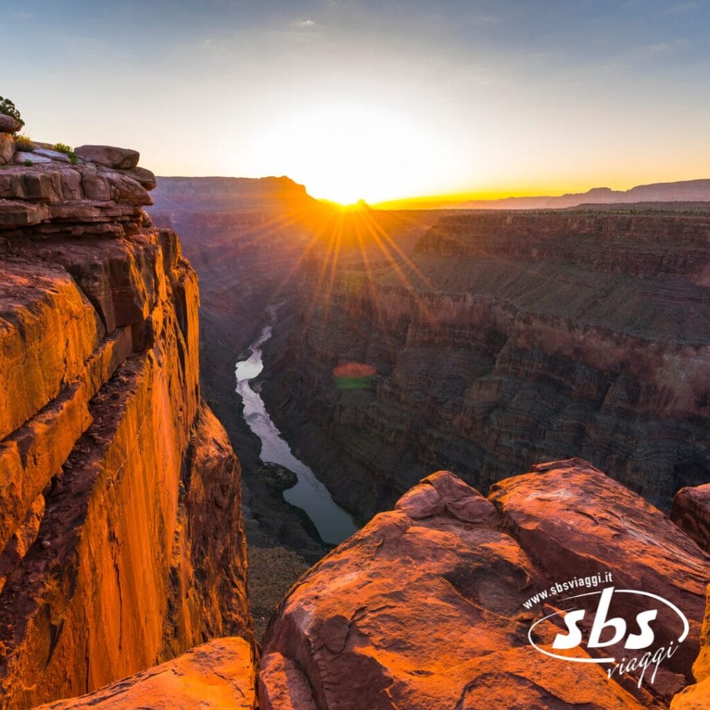Tramonto sul Grand Canyon con la luce del sole che illumina le scogliere rocciose arancioni e un fiume che serpeggia attraverso il canyon sottostante: lo scenario perfetto per qualsiasi Explorer 1924. Il logo
