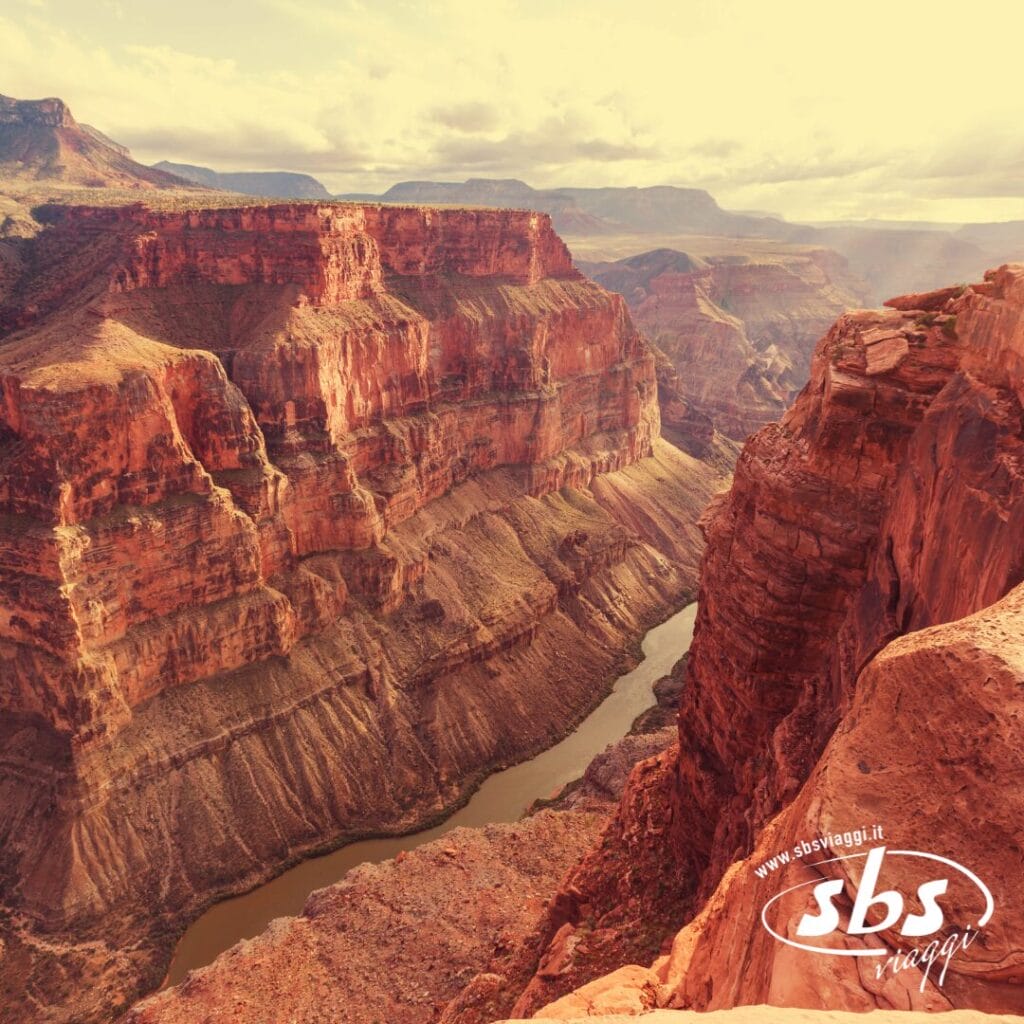 Un'ampia visuale del Grand Canyon mostra scogliere di roccia rossa stratificate e un fiume che serpeggia attraverso il fondo del canyon sotto un cielo limpido, che ricorda i paesaggi di Los Angeles immortalati nel 1924.