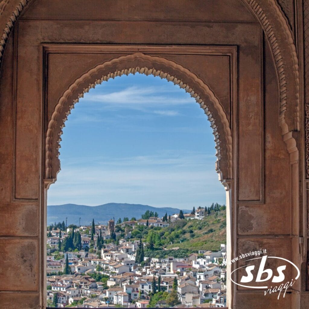 Vista attraverso un arco decorato che mostra una pittoresca cittadina andalusa con edifici bianchi, alberi verdi e dolci colline sotto un cielo azzurro: un pittoresco invito a visitare questa incantevole regione.