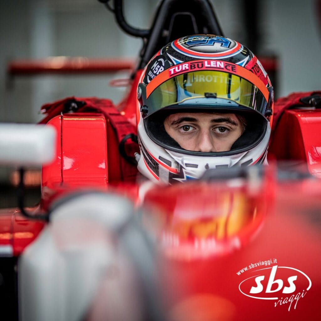 Un pilota con il casco è seduto a bordo di una monoposto rossa di Formula 1, con lo sguardo rivolto al futuro. L'auto presenta il logo