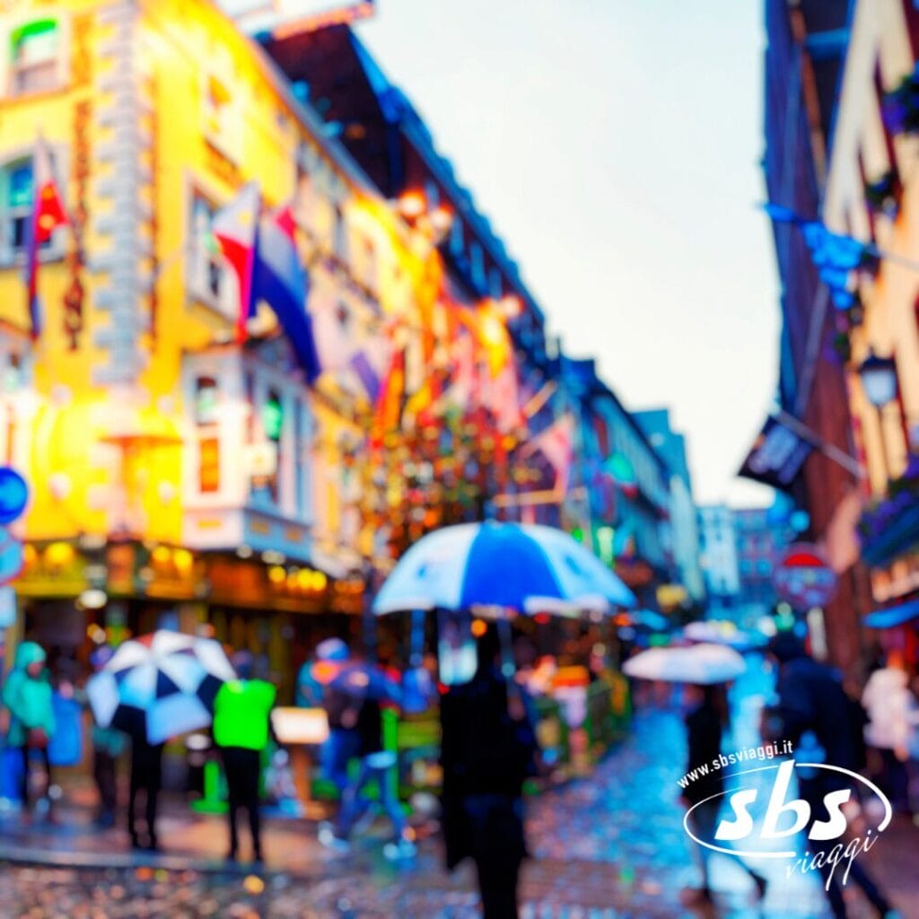 Scena di strada cittadina affollata in una giornata piovosa con persone che tengono in mano un ombrello; edifici colorati e luci natalizie sullo sfondo suggeriscono una serata perfetta per gustare un cocktail irlandese dopo aver seguito la vostra ricetta preferita.
