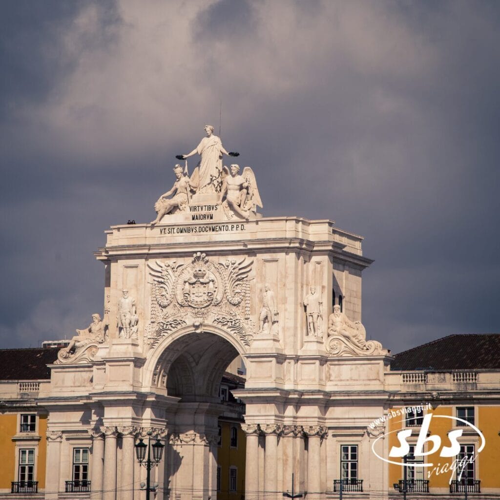 Un arco di trionfo in pietra adornato da statue si erge contro un cielo nuvoloso, con parte di un edificio giallo visibile dietro di esso: uno spettacolo iconico di qualsiasi Tour del Portogallo.