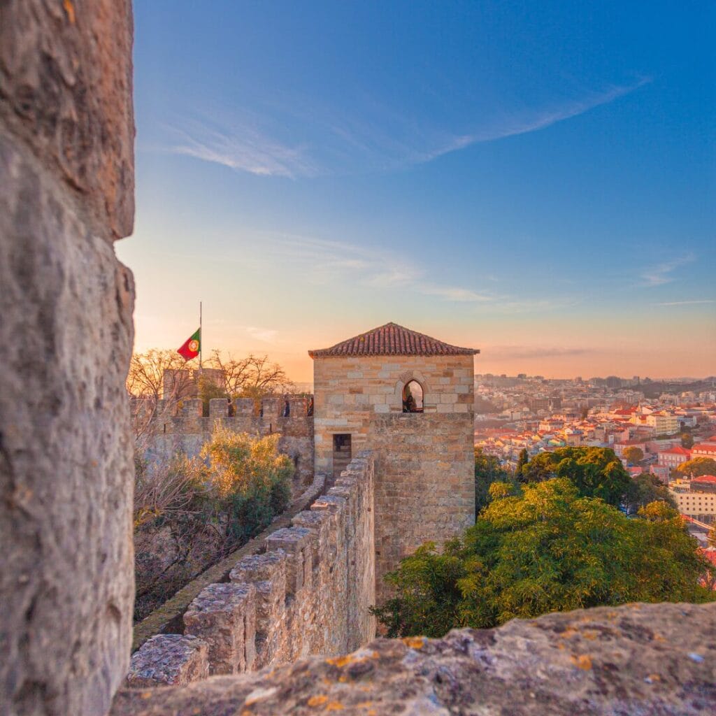 Le mura di pietra del castello e una torretta con una bandiera portoghese a mezz'asta dominano il paesaggio urbano al tramonto: uno scenario perfetto per iniziare il tuo tour del Portogallo o per sorseggiare un classico cocktail portoghese sotto il limpido cielo azzurro.