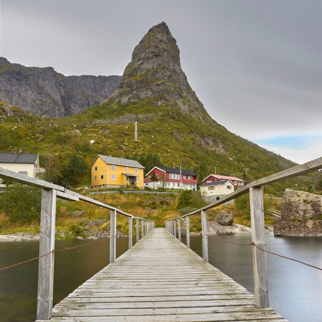Una passerella di legno conduce a un piccolo gruppo di case colorate ai piedi di una ripida montagna rocciosa, sotto un cielo coperto, nell'incantevole Terra dei Fiordi, in Norvegia.