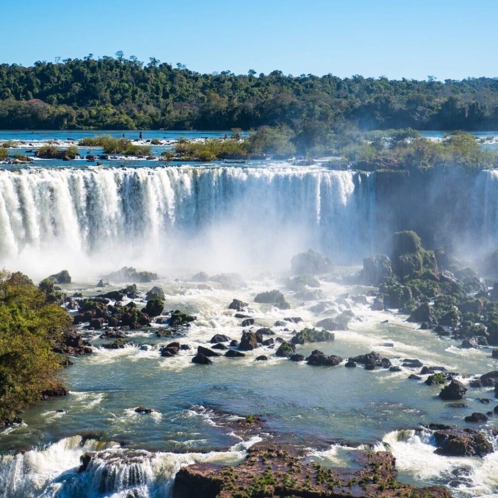 Un'ampia cascata si riversa da una parete rocciosa nel fiume sottostante, mentre la nebbia si alza e sullo sfondo si estende una fitta foresta sudamericana, sotto un cielo limpido.