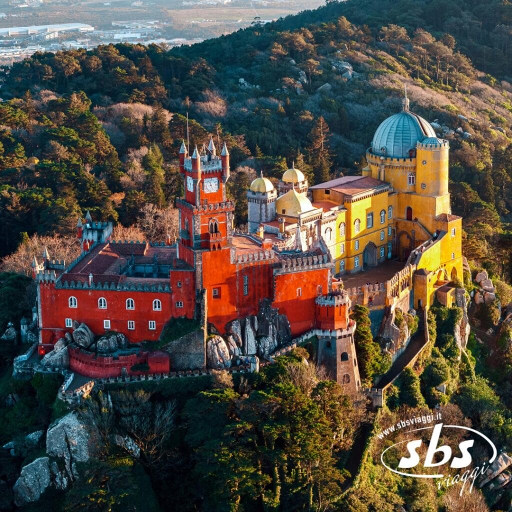 Vista aerea de Le Meraviglie del Portogallo: un palazzo colorato con sezioni rosse e gialle incastonato in una collina boscosa. La scena offre uno scorcio invitante della bellezza del Portogallo, con una pittoresca cittadina sullo sfondo, perfetta per il tuo prossimo tour.