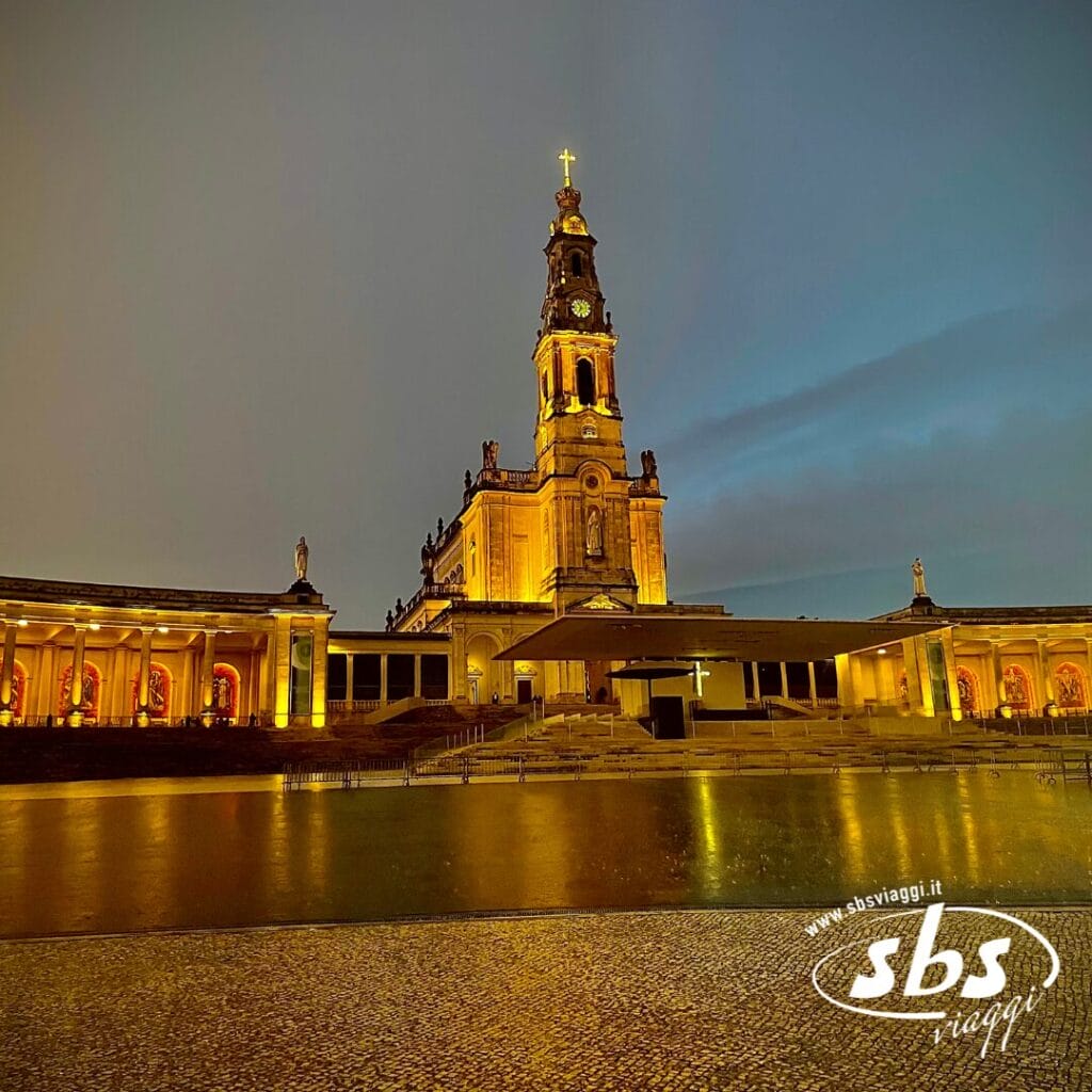 La chiesa illuminata, un faro nella notte portoghese, vanta un'alta torre dell'orologio sormontata da una croce. Strutture ad arco la circondano sotto il cielo crepuscolare, mentre la piazza acciottolata in primo piano invita a un tour veloce di bellezza senza tempo.