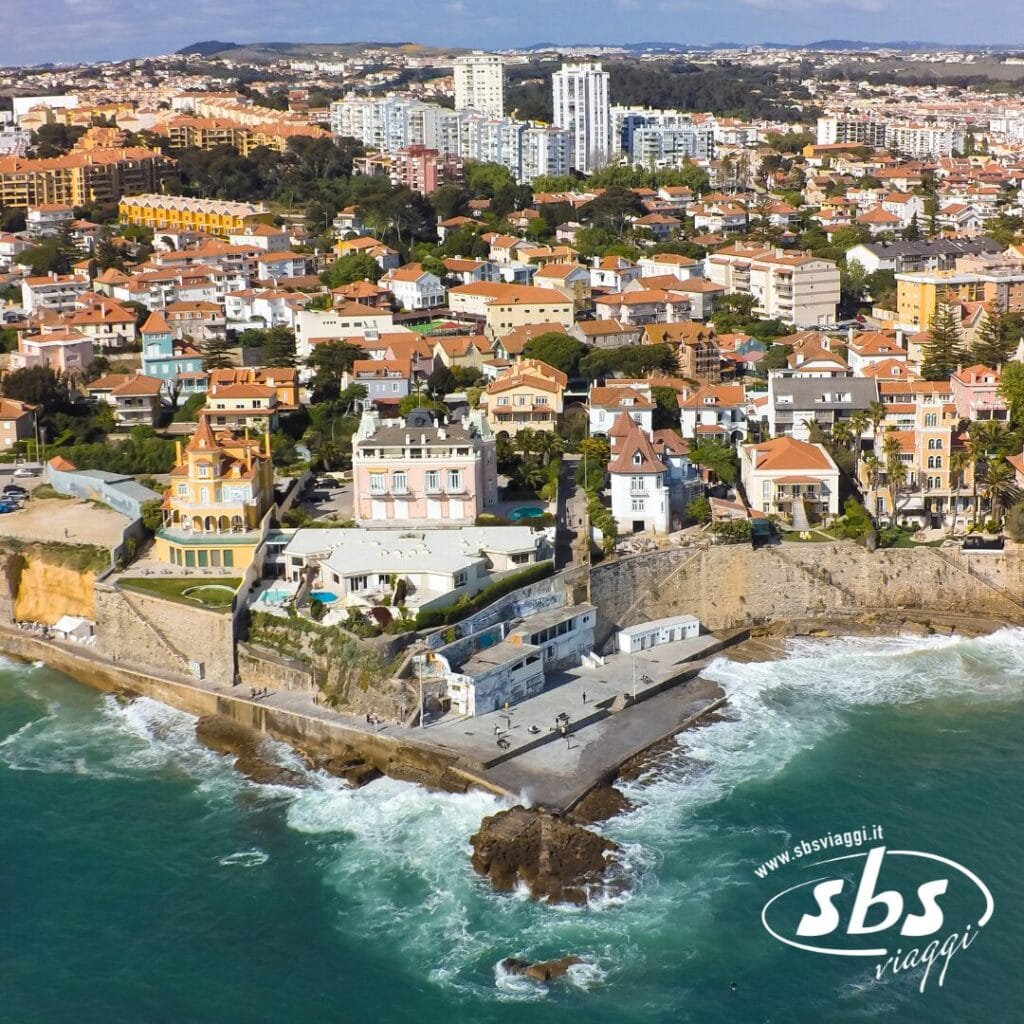 Vista aerea di una città costiera in Portogallo, con edifici vicino alla riva, onde che si infrangono contro una zona rocciosa e un paesaggio urbano sullo sfondo.