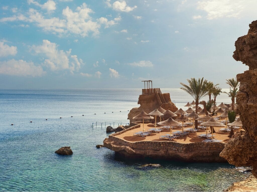 Una tranquilla vista costiera presenta acque azzurre e limpide, maestose formazioni rocciose e una spiaggia sabbiosa adornata da ombrelloni e lettini. Le palme costeggiano con grazia la costa sotto un cielo parzialmente nuvoloso, invitando al relax e ai momenti sereni tipici dei paesaggi di Bozza automatica.