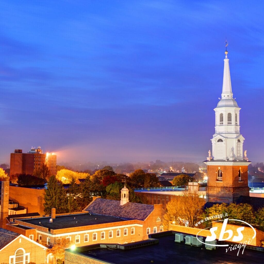 Il campanile bianco di una chiesa si erge sopra edifici in mattoni e alberi al crepuscolo, con un cielo nebbioso e le luci della città sullo sfondo, catturando il fascino di Metropoli durante il tuo tour della costa orientale.