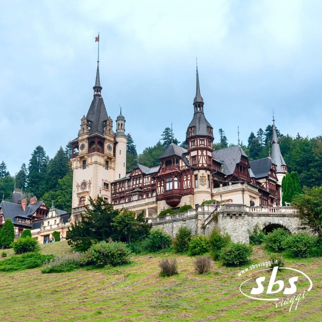 Il Castello di Peles, con le sue torri ornate, sorge su un pendio erboso in Romania, circondato da alberi sotto un cielo nuvoloso. Il logo