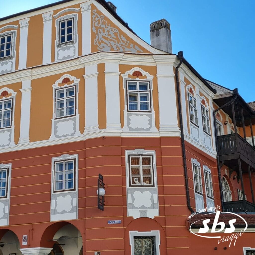 Un edificio storico di tre piani con dettagli decorativi arancioni e bianchi, numerose finestre e un balcone in legno sul lato destro. Il logo