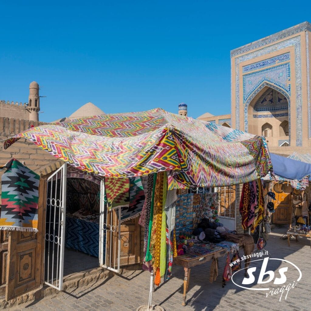 Una vivace bancarella di mercato con tessuti e stoffe dai motivi colorati provenienti da Via Della Seta è esposta sotto una tettoia, di fronte a un'architettura storica con piastrelle blu in una giornata di sole.