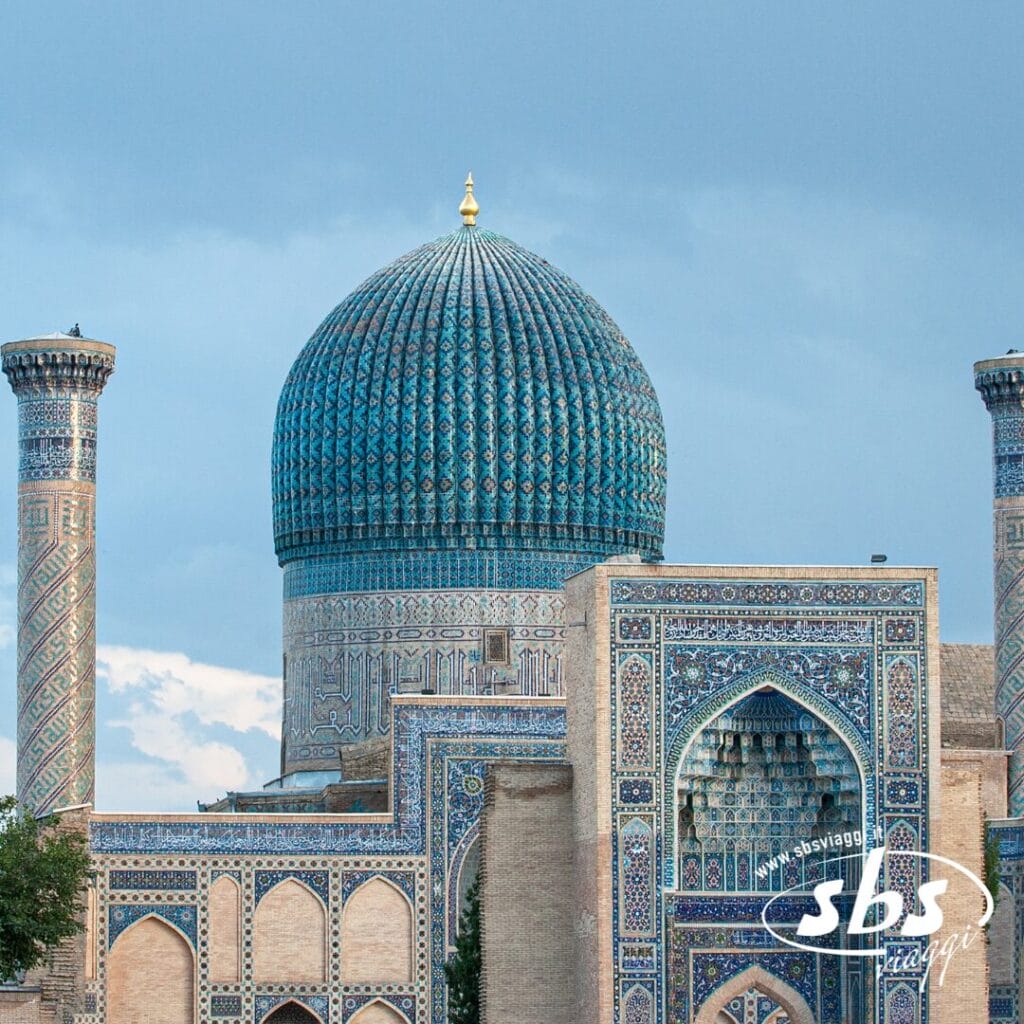 Una grande struttura a cupola rivestita di piastrelle blu, con intricati motivi a mosaico e alti minareti si erge maestosa contro un cielo parzialmente nuvoloso, evocando la grandiosità della Valle di Fergana nella storica Terra di Tamerlano.