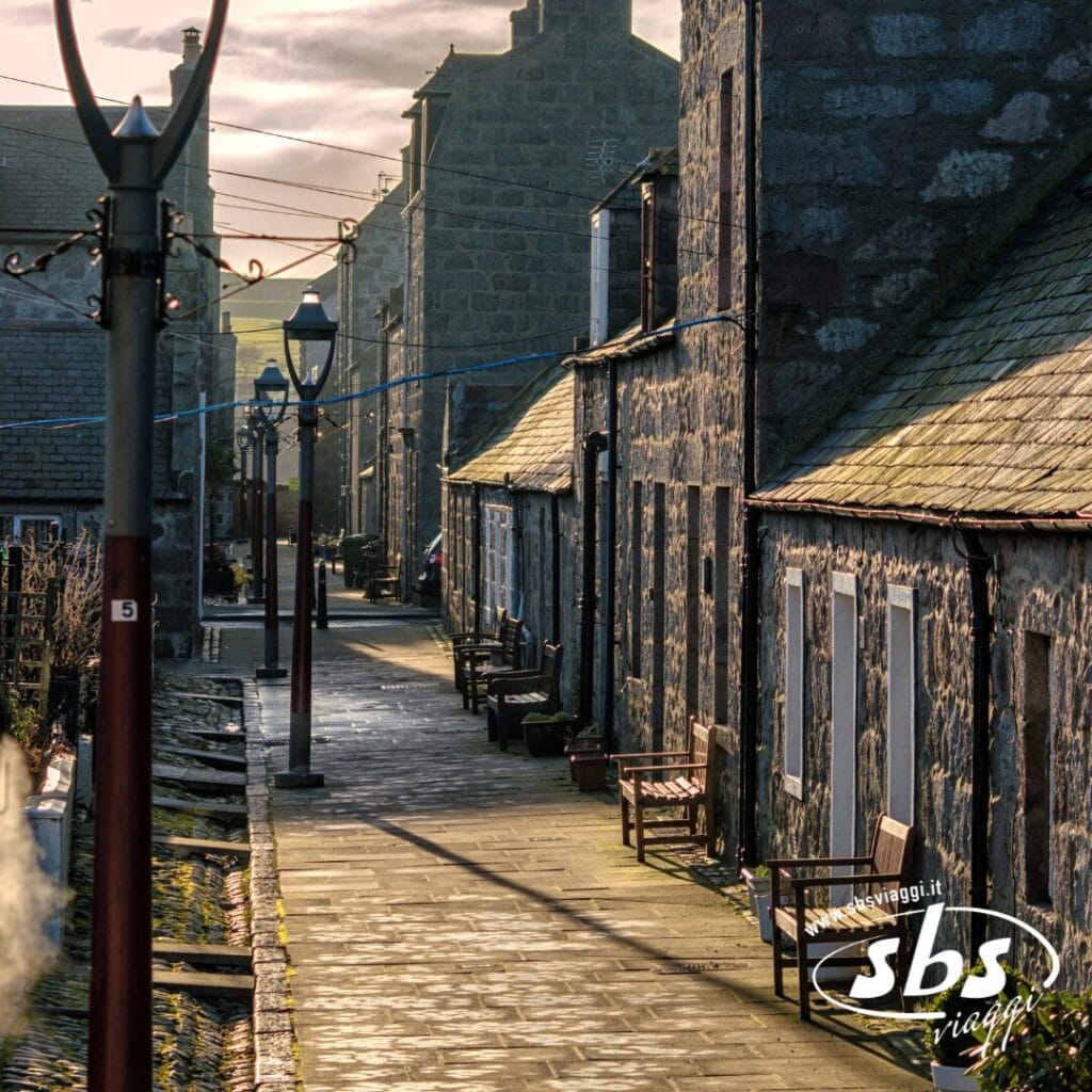 Una stretta strada lastricata di pietra fiancheggiata da vecchi edifici in pietra e panchine, illuminata dalla tenue luce del sole: perfetta per chi cerca il fascino di un viaggio in Scozia.