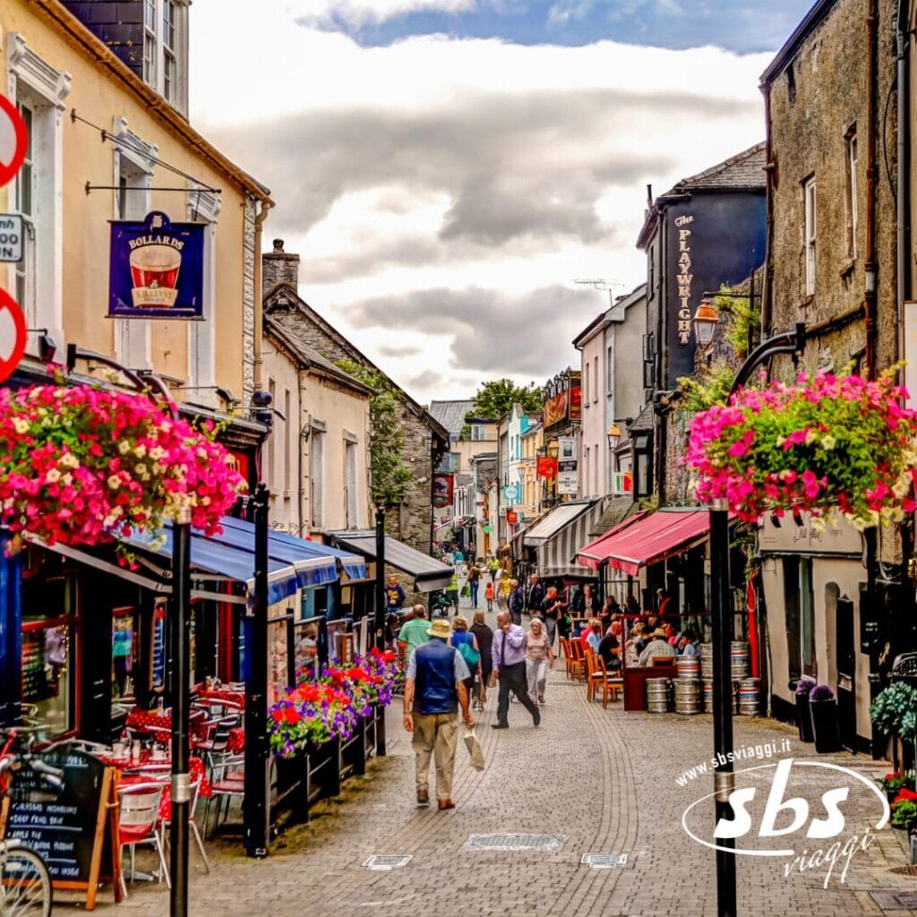 Una strada acciottolata fiancheggiata da negozi, ristoranti e cesti di fiori appesi; la gente passeggia nell'area pedonale sotto Cieli d'Irlanda, l'incantevole cielo irlandese.