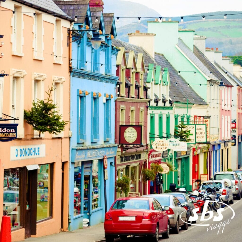 Una fila di edifici colorati con negozi e attività commerciali costeggia una stretta strada con auto parcheggiate, catturando il fascino di una piccola città durante un viaggio attraverso le bellezze d'Irlanda.
