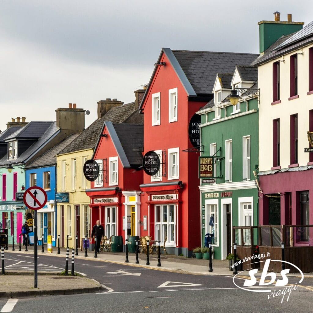 Una fila di edifici colorati, tra cui negozi e un hotel, costeggia l'angolo di una strada di una piccola cittadina irlandese, con un cielo coperto e pedoni nelle vicinanze, evocando il fascino di luoghi come le Isole Aran.