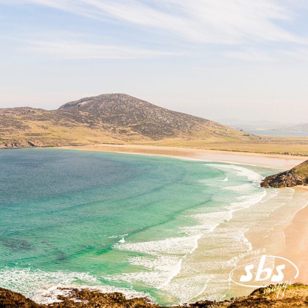 Veduta di una spiaggia sabbiosa curva vicino a Belfast, Irlanda, con acqua turchese, onde dolci e colline erbose sullo sfondo sotto un cielo parzialmente nuvoloso.