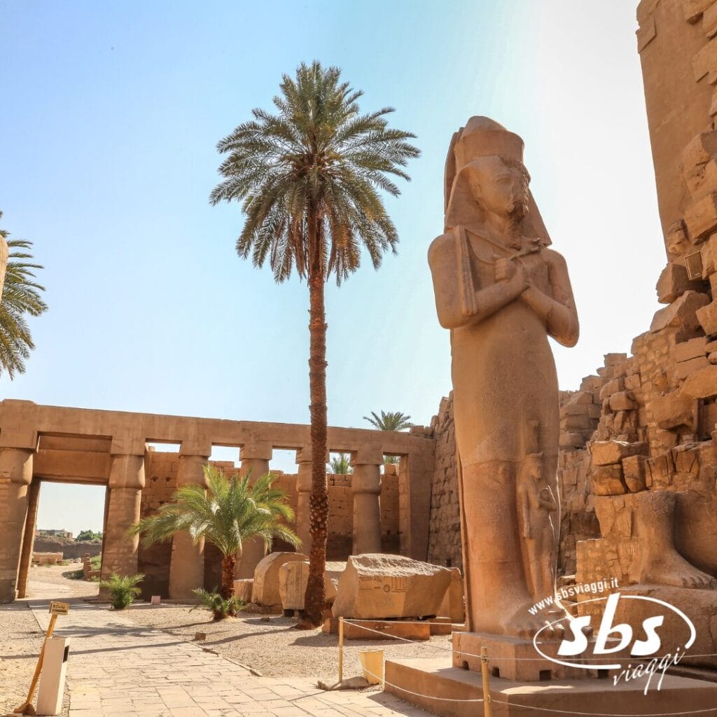Statua dell'antico Egitto e palme in un sito archeologico vicino al Cairo, con rovine in pietra e un ingresso sotto un cielo soleggiato.