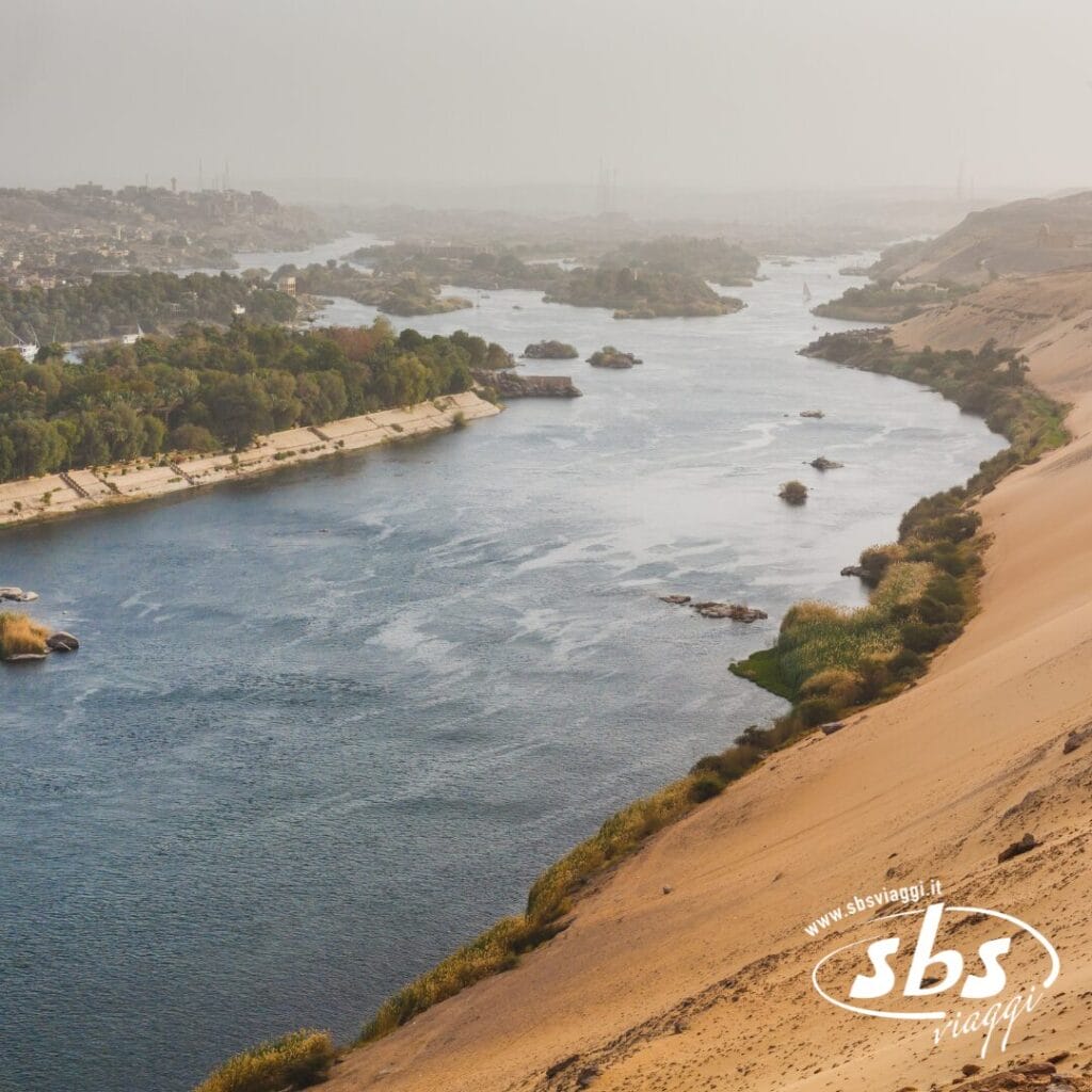 Un ampio fiume, il Nilo, scorre attraverso un paesaggio desertico con rive sabbiose e macchie di vegetazione verde lungo la riva.