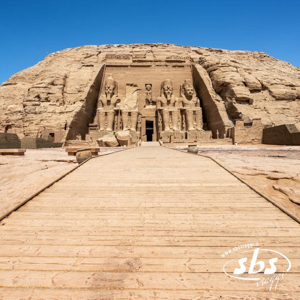 Un'ampia passerella in legno conduce all'antico tempio di Abu Simbel nell'Antico Egitto, caratterizzato da grandi statue sedute scolpite in una scogliera di arenaria sotto un cielo azzurro e limpido: un luogo iconico spesso ammirato durante una Crociera sul Nilo.