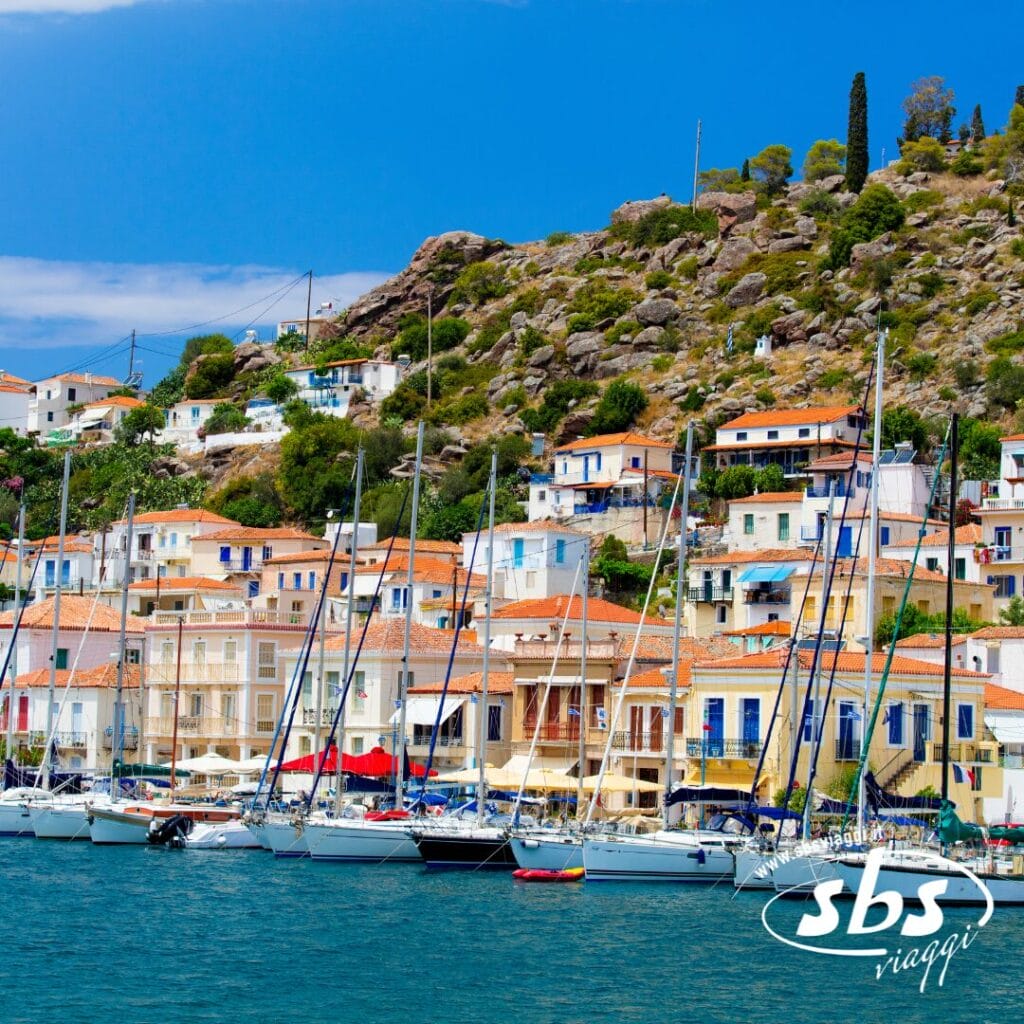 Case colorate con tetti arancioni fiancheggiano una collina dietro un porto turistico pieno di barche a vela, catturando il fascino di un'avventura del Tour Grecia. Il logo di SBS Viaggi è visibile nell'angolo in basso a destra.